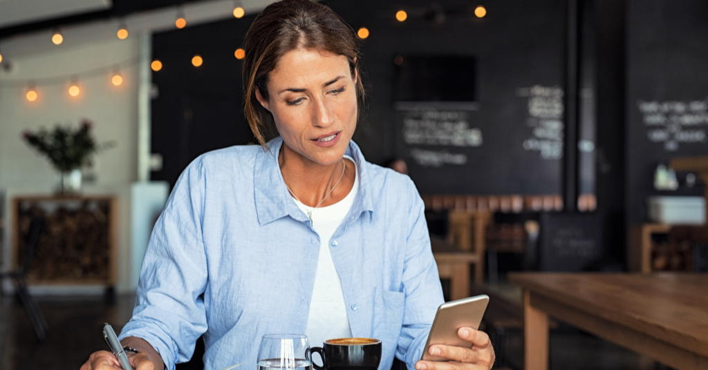 Mature woman sitting in cafe at table and typing a message on smartphone. Middle aged blogger making notes using organizing application on phone. Businesswoman reading information from smartphone while working remotely in cafe interior.